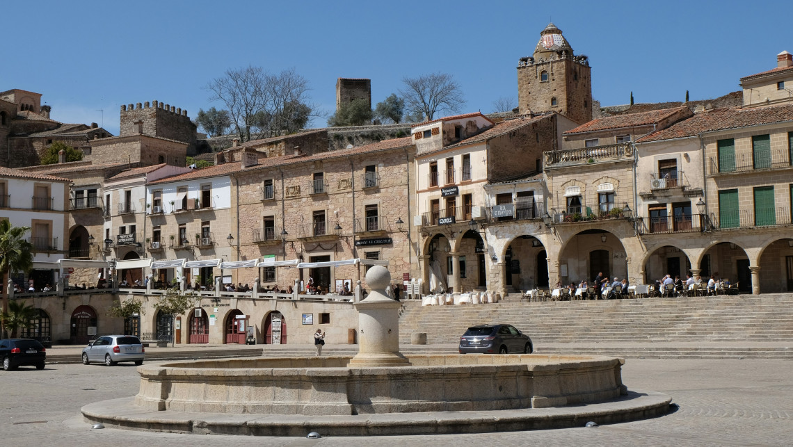 A stone fountain stands in Plaza Mayor in Trujillo, C&aacute;ceres, Spain, surrounded by broad steps, caf&eacute; terraces and arcaded historic buildings. Medieval towers rise above the rooftops under a clear blue sky.