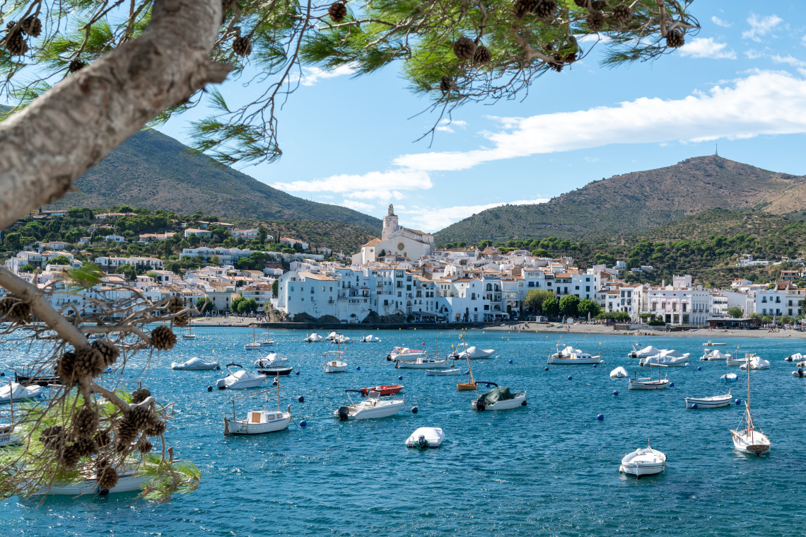 Boats are anchored across the blue harbour of Cadaqu&eacute;s on the Costa Brava in Spain. The town&rsquo;s white waterfront buildings extend along the bay beneath Santa Maria Church and the hills of Cap de Creus.