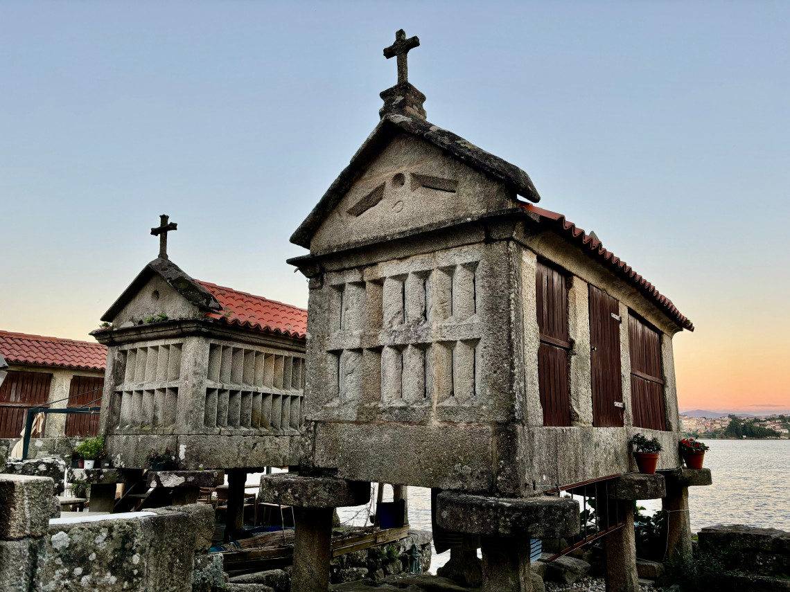 Two traditional stone h&oacute;rreos stand on raised pillars beside the water in Combarro, Pontevedra, Spain. Crosses crown the granite granaries, which feature wooden side panels and red-tiled roofs.