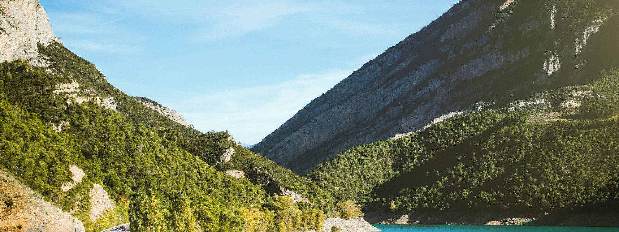 Forested limestone mountains surround the waters of Canelles Reservoir at Mont-rebei Gorge in Lleida, Spain. Layered rock formations rise above the narrow valley beneath a clear sky.