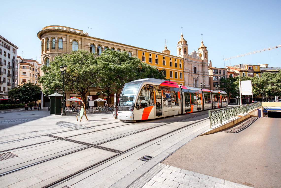 A modern red-and-silver tram travels through the historic centre of Zaragoza, Spain. Warm-toned buildings, mature trees and a twin-towered church frame the urban scene.