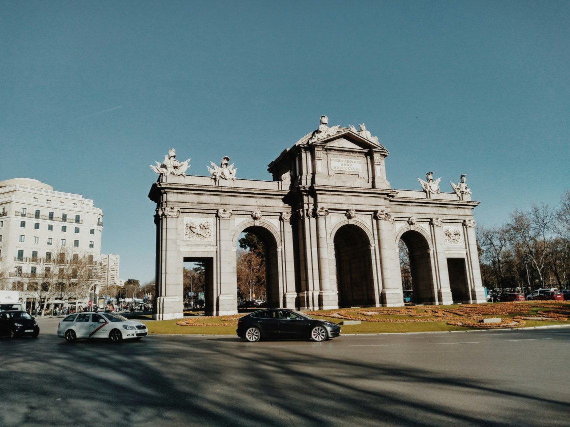 The Puerta de Alcal&aacute; stands at Plaza de la Independencia in Madrid, Spain, beneath a clear blue sky. Cars pass around the stone monument, which features five openings and sculpted decorative details.
