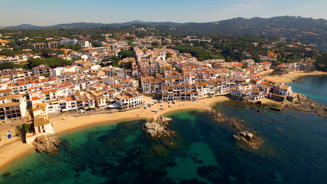 Aerial view of Calella de Palafrugell, Spain, showing whitewashed buildings and terracotta rooftops beside sandy beaches and rocky coves. Clear Mediterranean water surrounds the coastal village on the Costa Brava.
