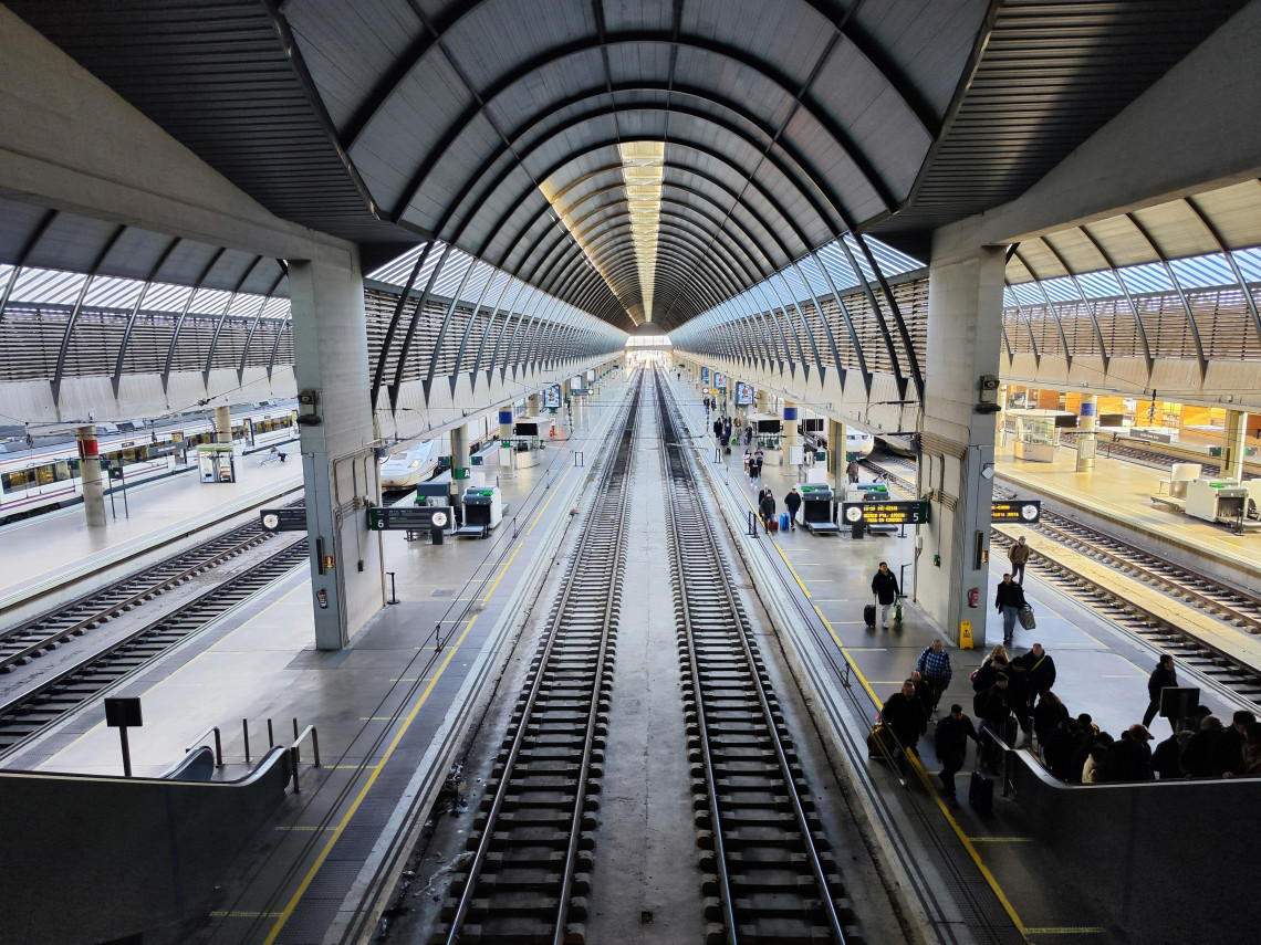 Elevated view across the platforms and railway tracks inside Seville-Santa Justa Railway Station in Seville, Spain. Passengers wait beneath the station&rsquo;s long vaulted roof while escalators connect the platforms to the upper level.