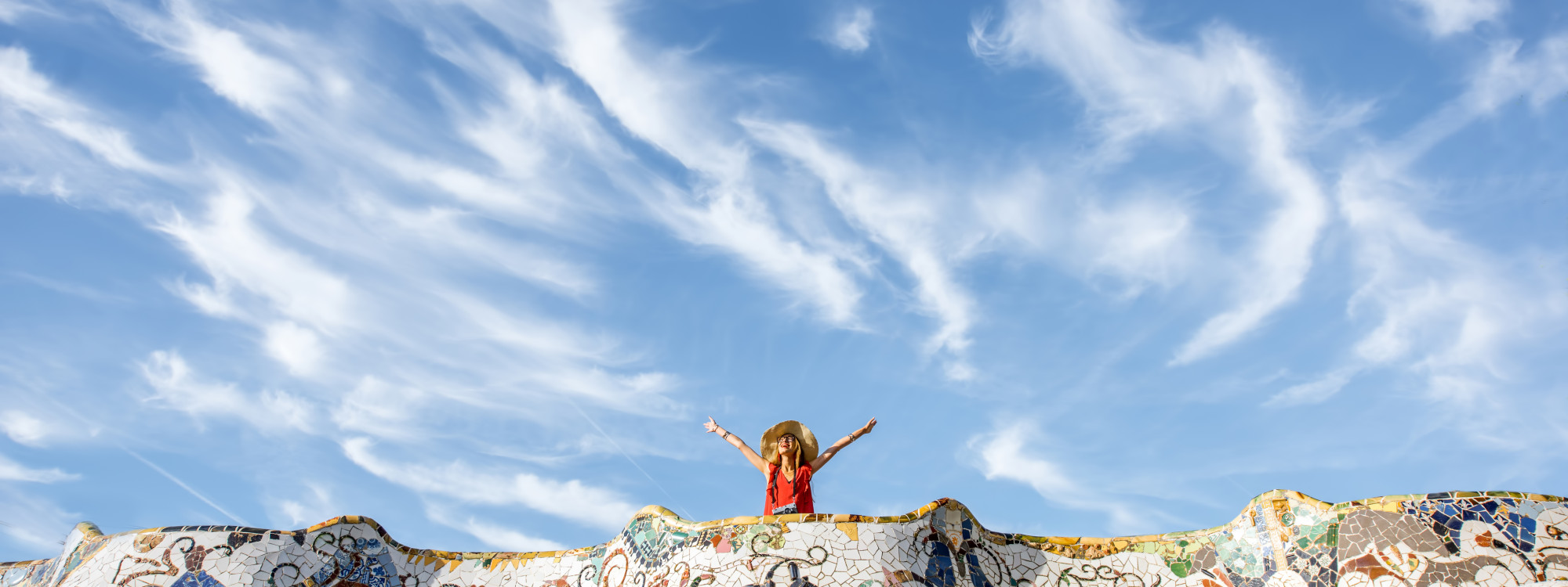 A visitor stands behind the colourful mosaic terrace at Park Güell in Barcelona, Spain, with arms raised beneath a wide blue sky. The curved trencadís surface frames the lower part of the scene.