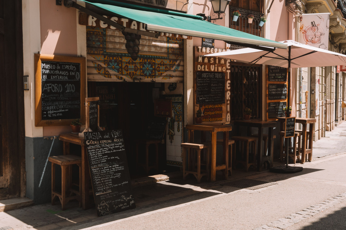 The exterior of Mas&iacute;a del Vino in Valencia, Spain, features a green awning, wooden stools and handwritten menus beside the entrance. Decorative tiles and chalkboards reflect the traditional character of the tavern.