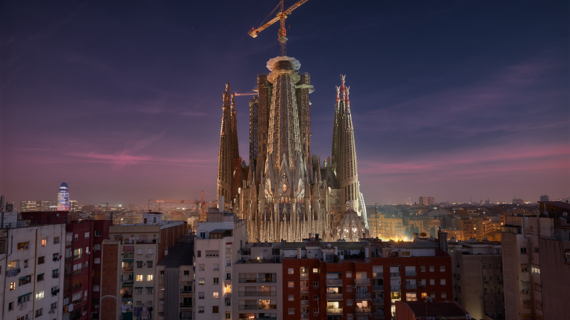 The Sagrada Fam&iacute;lia rises above residential buildings in Barcelona, Spain, illuminated against a purple and pink evening sky. Construction cranes and the basilica&rsquo;s sculpted towers form part of the city skyline.