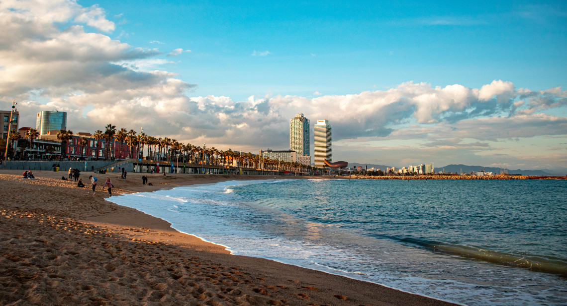 Barceloneta Beach in Barcelona, Spain, curves along the Mediterranean coast beneath a partly cloudy sky. People walk on the sand while palm trees and modern waterfront towers rise beside the promenade.