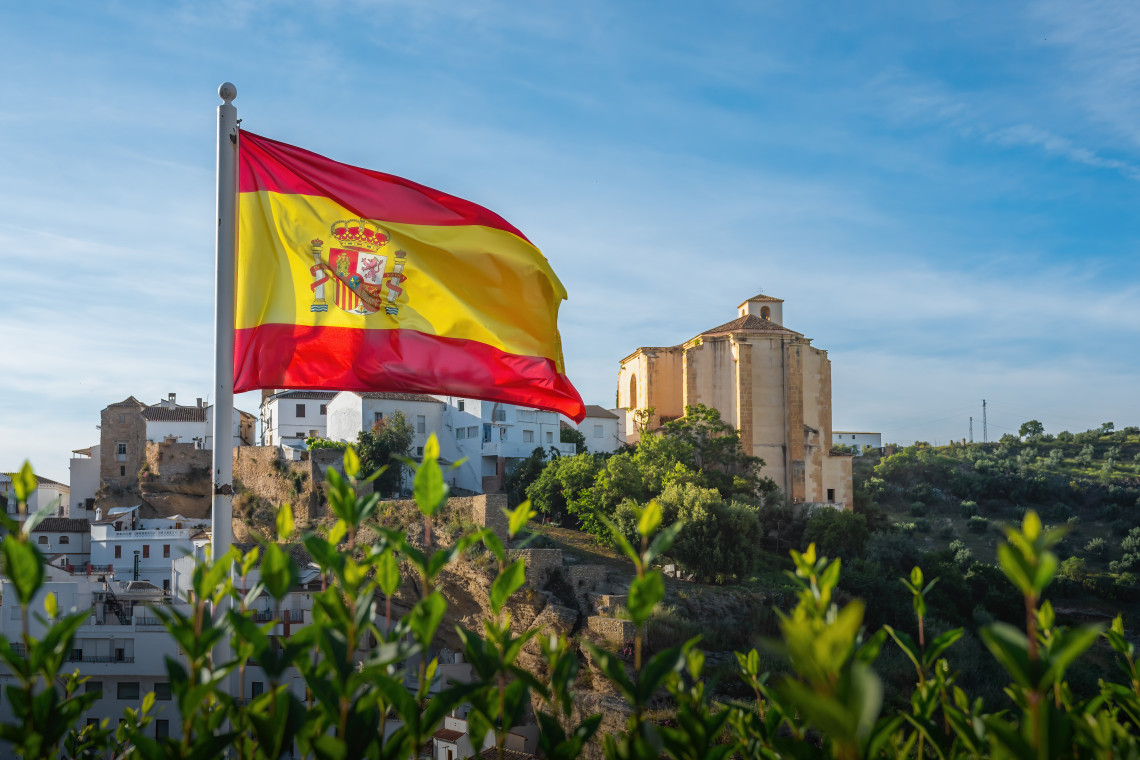 A Spanish flag waves above the whitewashed town of Setenil de las Bodegas in C&aacute;diz, Spain. The Church of Nuestra Se&ntilde;ora de la Encarnaci&oacute;n and historic buildings rise across the hillside behind green foliage.