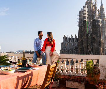 A couple enjoys a meal on a rooftop terrace overlooking the Sagrada Família in Barcelona, Spain. Plates of food and drinks are arranged on the table, with the basilica’s towers rising behind them.