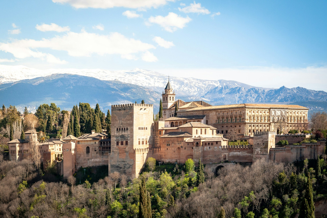 Panoramic view of the Alhambra in Granada, Spain, with its fortified towers and palace buildings rising above a wooded hillside. The snow-covered Sierra Nevada mountains extend across the background.