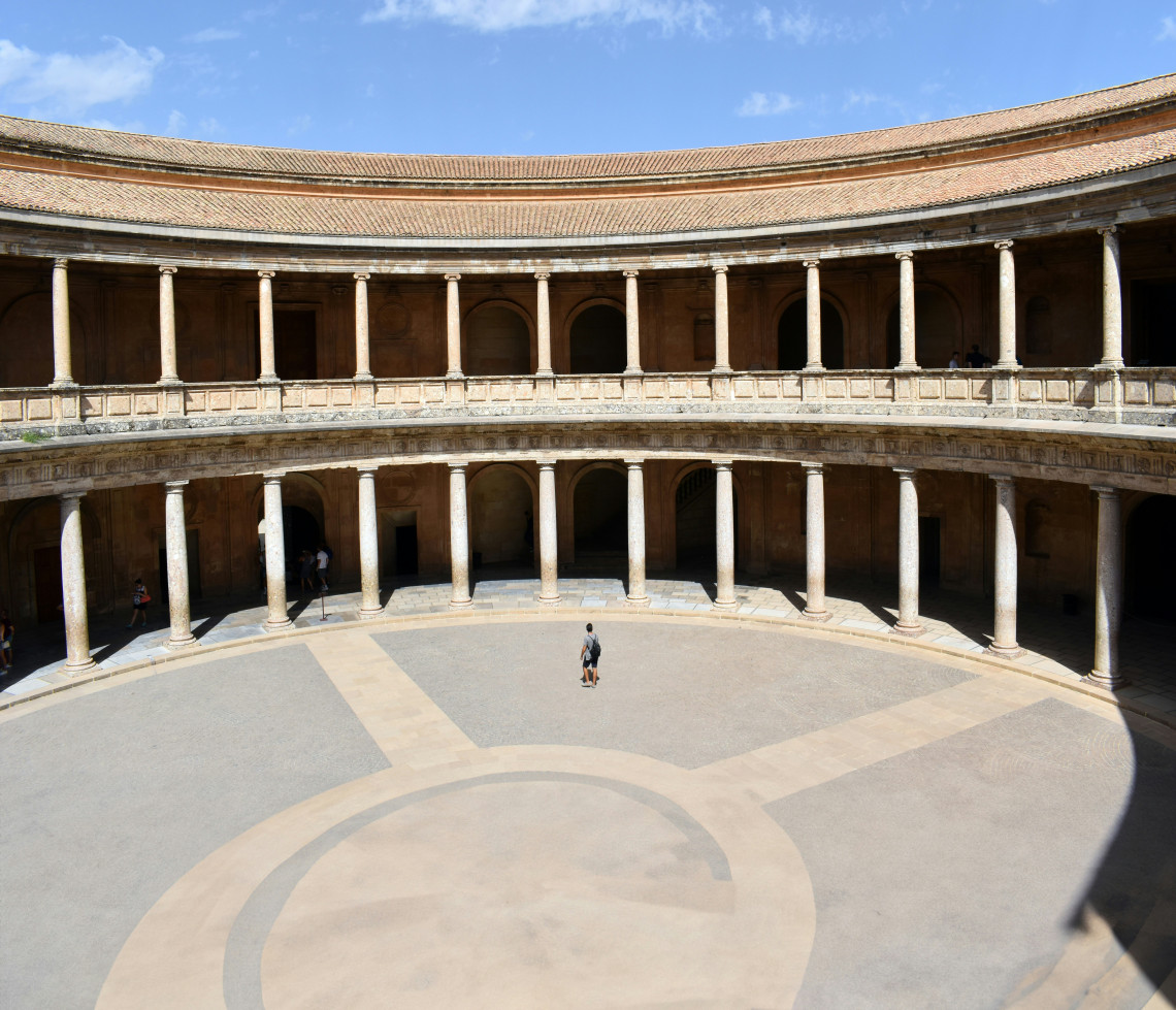 The circular courtyard of the Palace of Charles V at the Alhambra in Granada, Spain, is surrounded by two levels of stone columns. Visitors cross the open Renaissance courtyard beneath a clear blue sky.