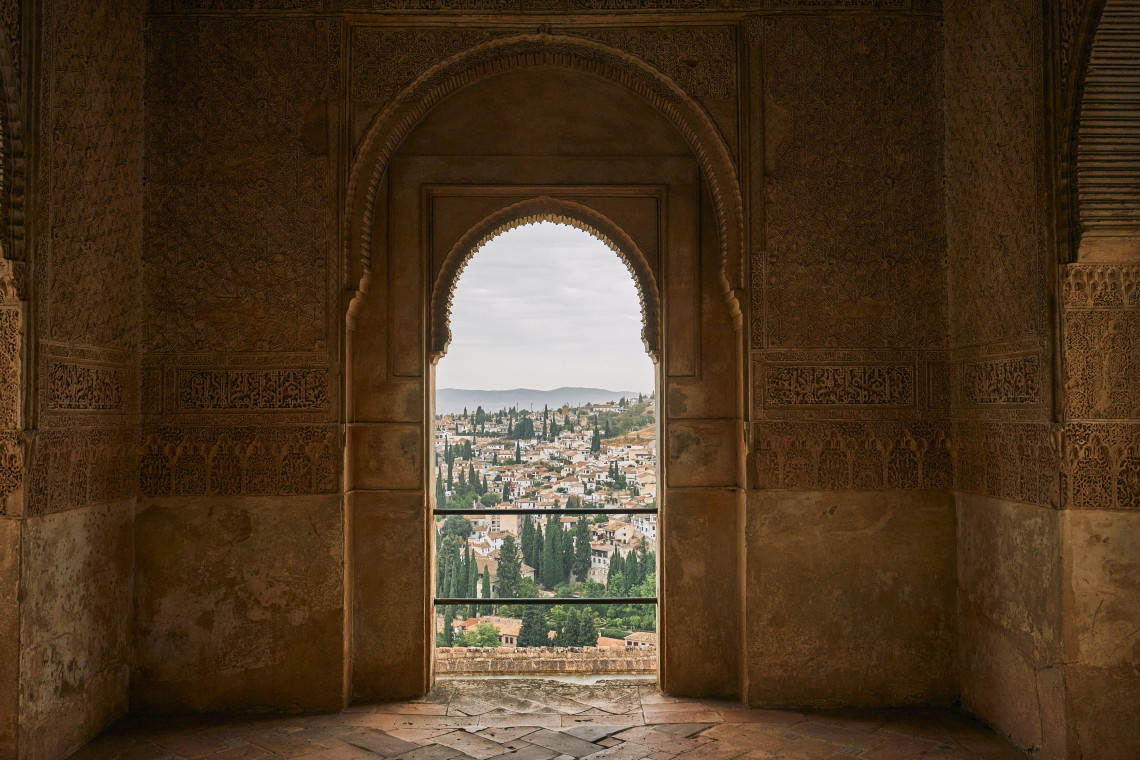 The historic Albaicín district of Granada, Spain, is visible through a carved Moorish arch inside the Alhambra. White buildings, cypress trees and surrounding hills extend beyond the palace opening.