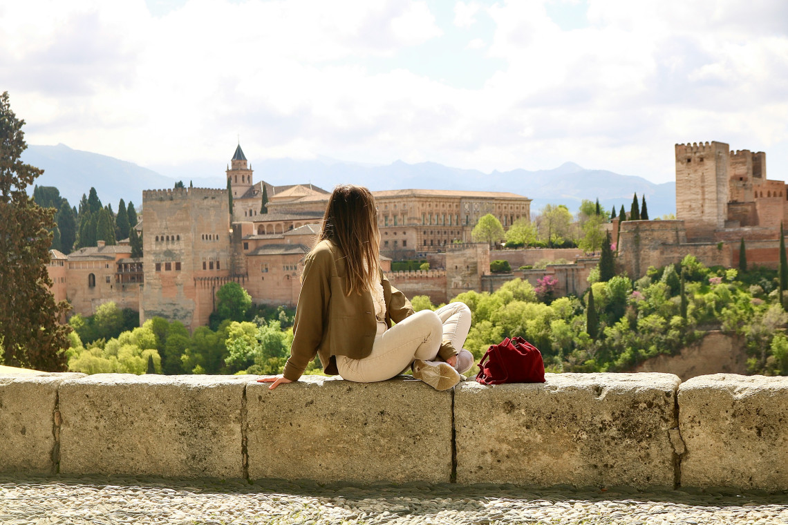 A traveller sits on a stone wall at Mirador de San Nicolás in Granada, Spain, looking across towards the Alhambra. The palace complex, green hillside and Sierra Nevada mountains fill the background.
