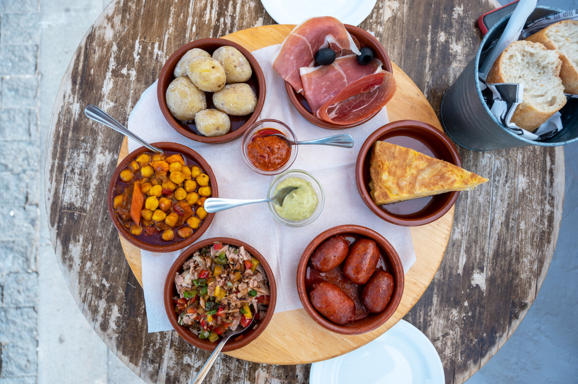 A selection of Spanish tapas is arranged on an outdoor table in Granada, Spain. The meal includes tortilla, chorizo, cured ham, potatoes, chickpeas, bread and small bowls of sauces.