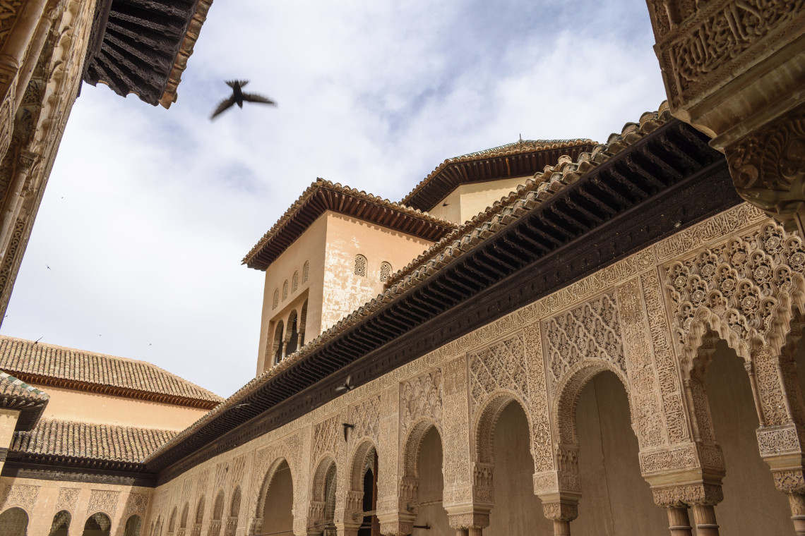 Moorish arches and carved stucco decorate the Nasrid Palaces inside the Alhambra in Granada, Spain. Detailed geometric patterns, tiled roofs and a historic tower frame the open courtyard.