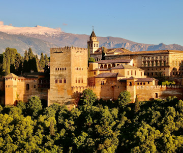 The Alhambra rises above the wooded hills of Granada, Spain, with the snow-covered Sierra Nevada mountains in the background. Medieval towers, palace buildings and terracotta rooftops form the historic complex.
