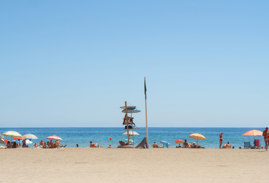 Visitors relax beneath colourful umbrellas on the wide sandy shore of La Malagueta Beach in M&aacute;laga, Spain. A lifeguard station stands near the calm Mediterranean Sea beneath a clear blue sky.
