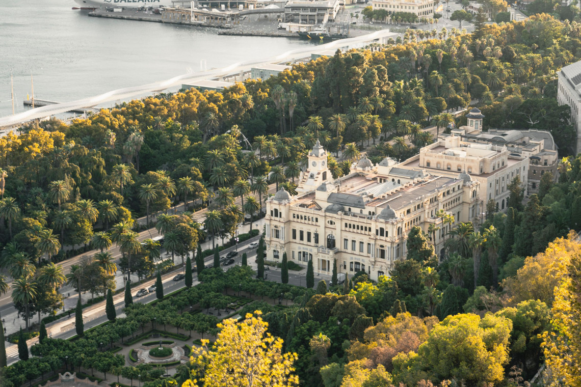 Aerial view of M&aacute;laga City Hall in M&aacute;laga, Spain, surrounded by the landscaped gardens and palm trees of Paseo del Parque. The city&rsquo;s port and waterfront are visible beyond the historic building.