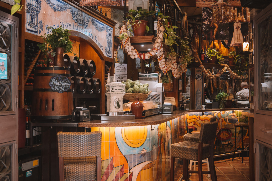 A traditional tapas bar in M&aacute;laga, Spain, features a colourful tiled counter, wooden stools and shelves filled with local products. Hanging garlic, decorative ceramics and rustic details create a characteristic Andalusian setting.