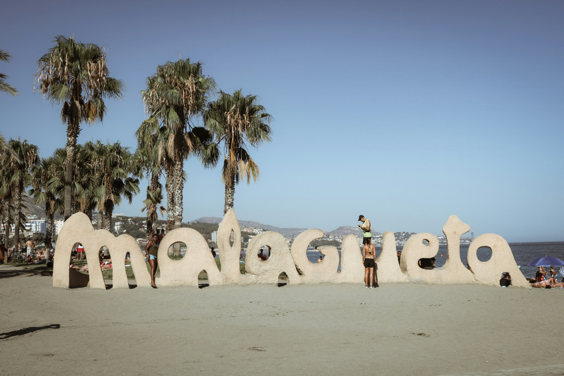 The sculptural La Malagueta sign stands on the beachfront in M&aacute;laga, Spain, surrounded by palm trees. Visitors gather around the landmark beside the Mediterranean Sea.