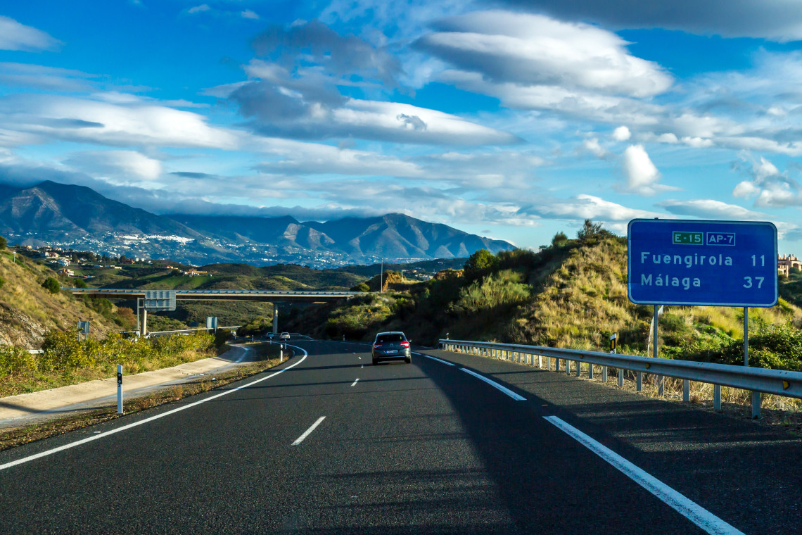 The AP-7 motorway passes through the mountainous landscape near Fuengirola, Spain, with a road sign directing travellers towards Fuengirola and M&aacute;laga. A vehicle follows the open road beneath layered clouds.