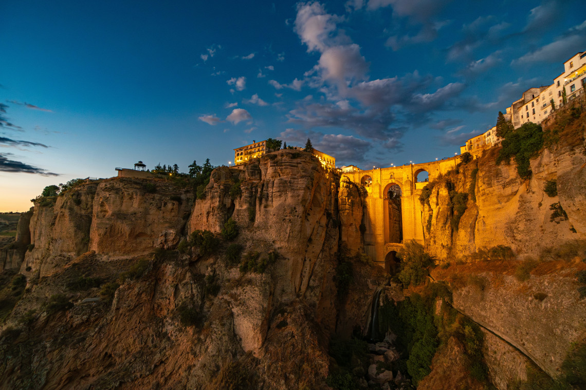 Puente Nuevo spans the deep El Tajo Gorge in Ronda, Spain, illuminated by warm evening light. Historic buildings rise above the rocky cliffs beneath a darkening blue sky.