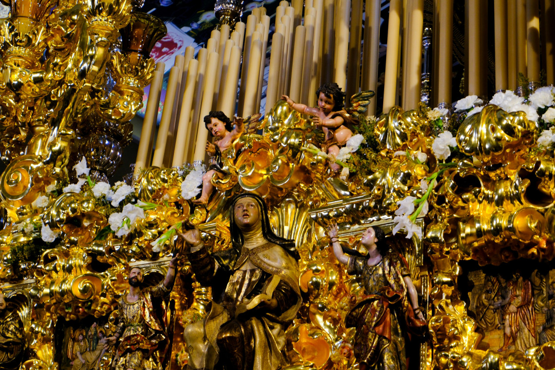 An elaborate gilded religious sculpture in M&aacute;laga, Spain, depicts a Carmelite saint surrounded by angels, floral details and carved ornamentation. Tall organ pipes form the backdrop to the intricate sacred artwork.