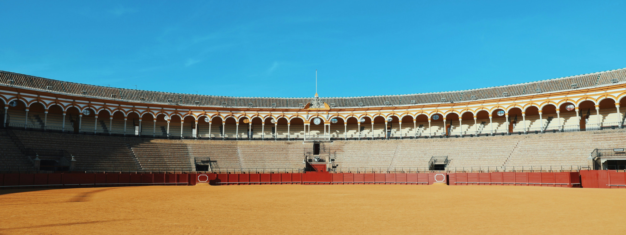 The interior of Plaza de Toros de la Maestranza in Seville, Spain, shows the sandy arena surrounded by shaded spectator seating and a continuous arched gallery.