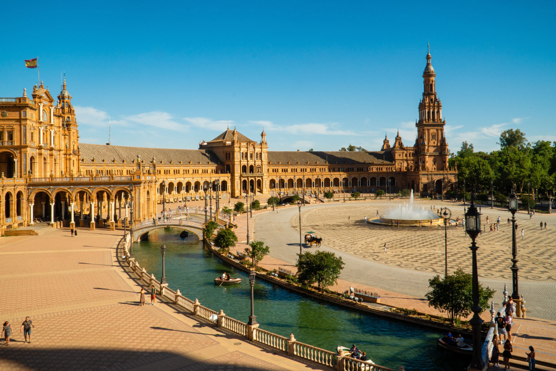 Panoramic view of Plaza de Espa&ntilde;a in Seville, Spain, showing its canal, decorative bridges and central fountain. The landmark&rsquo;s long arcaded fa&ccedil;ade and tall tower extend across the square.