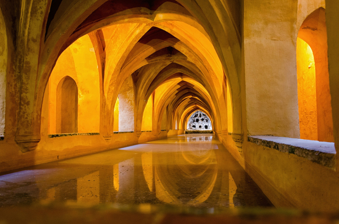 The Baths of Do&ntilde;a Mar&iacute;a de Padilla at the Real Alc&aacute;zar in Seville, Spain, feature a long reflective pool beneath repeating Gothic arches. Warm lighting highlights the vaulted underground passage.