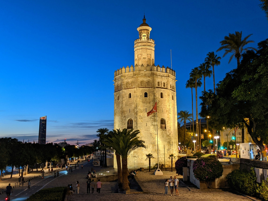 Torre del Oro stands illuminated beside the Guadalquivir River in Seville, Spain. Palm trees and pedestrians surround the historic stone tower beneath a deep blue evening sky.