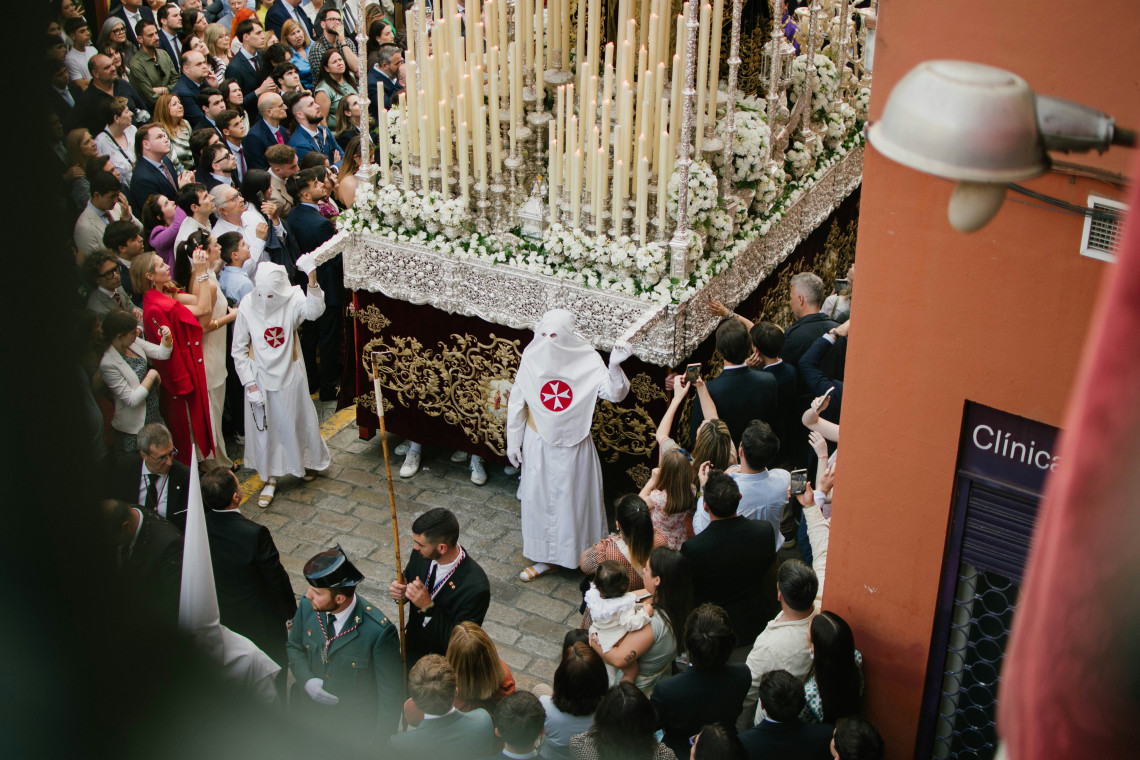 A Holy Week procession moves through a crowded street in Seville, Spain, with participants in traditional robes accompanying an ornate candlelit religious float. Spectators gather closely along both sides of the route.
