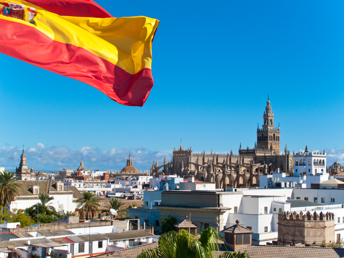 A Spanish flag waves above the rooftops of Seville, Spain, with Seville Cathedral and the La Giralda bell tower dominating the skyline. Whitewashed buildings, palm trees and historic towers fill the foreground.