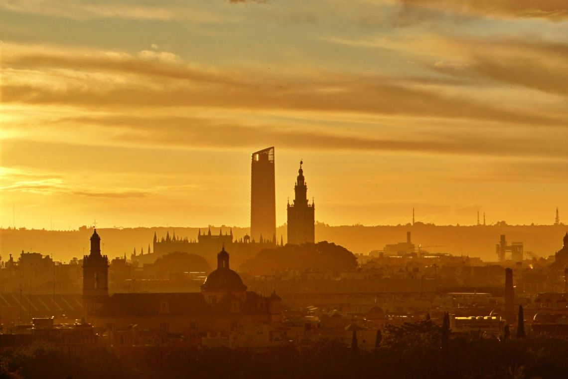 The skyline of Seville, Spain, appears in silhouette during a golden sunset. La Giralda rises near the modern Torre Sevilla, with historic rooftops and church domes filling the foreground.