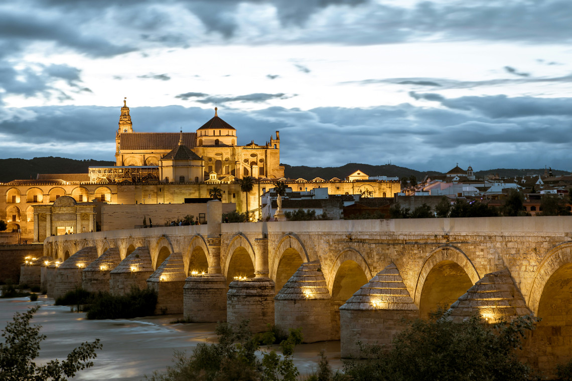 The illuminated Roman Bridge crosses the Guadalquivir River towards the Mosque-Cathedral of C&oacute;rdoba, Spain. Historic arches, stone walls and the cathedral tower stand beneath a cloudy evening sky.