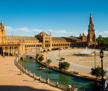Plaza de España in Seville, Spain, features a curved canal, tiled bridges and an expansive semicircular building. Ornate towers and covered galleries frame the open square beneath a clear blue sky.