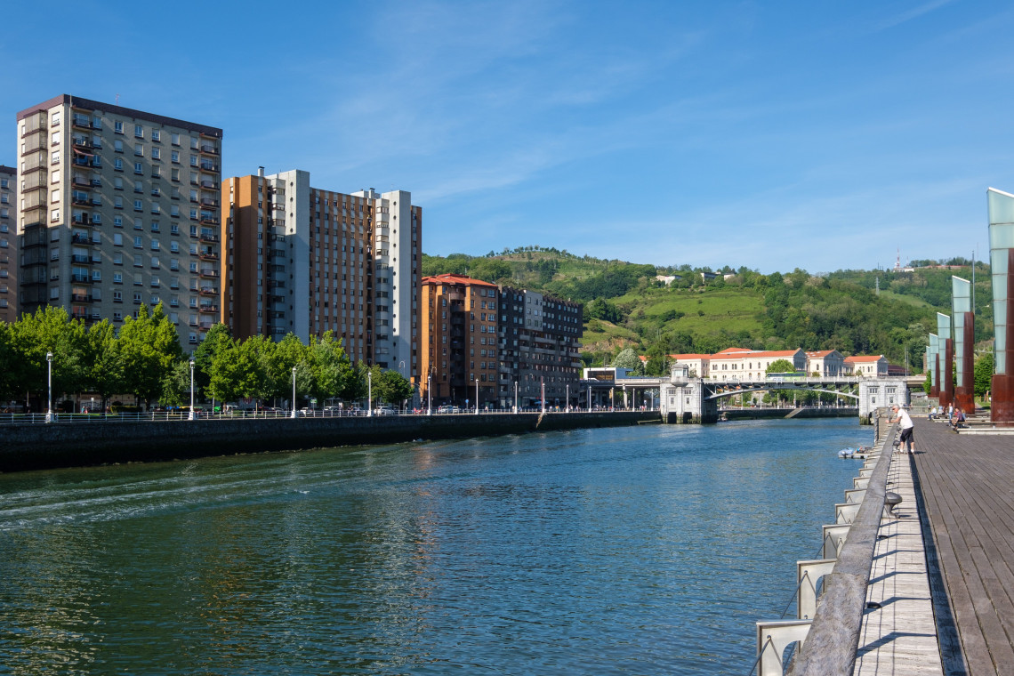 The Bilbao Estuary flows through Bilbao, Spain, beside apartment buildings, a wooden riverside promenade and a bridge framed by green hills.