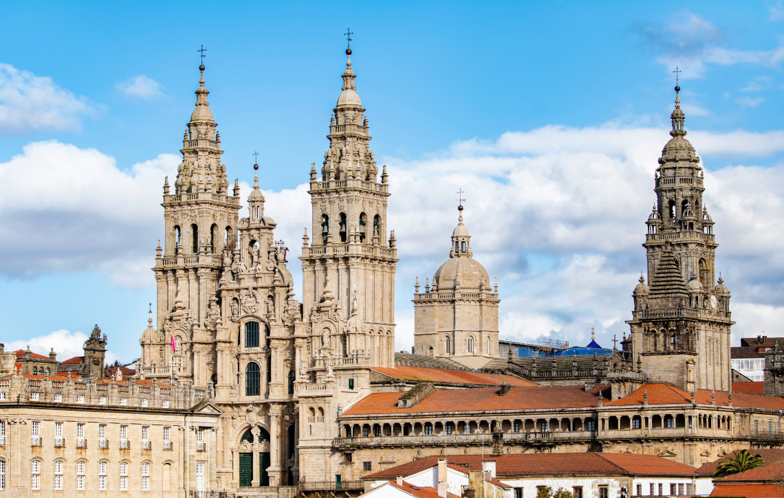 Santiago de Compostela Cathedral rises above the historic centre of Santiago de Compostela, Spain. Its carved stone towers, domes and terracotta rooftops stand beneath a blue sky with scattered clouds.