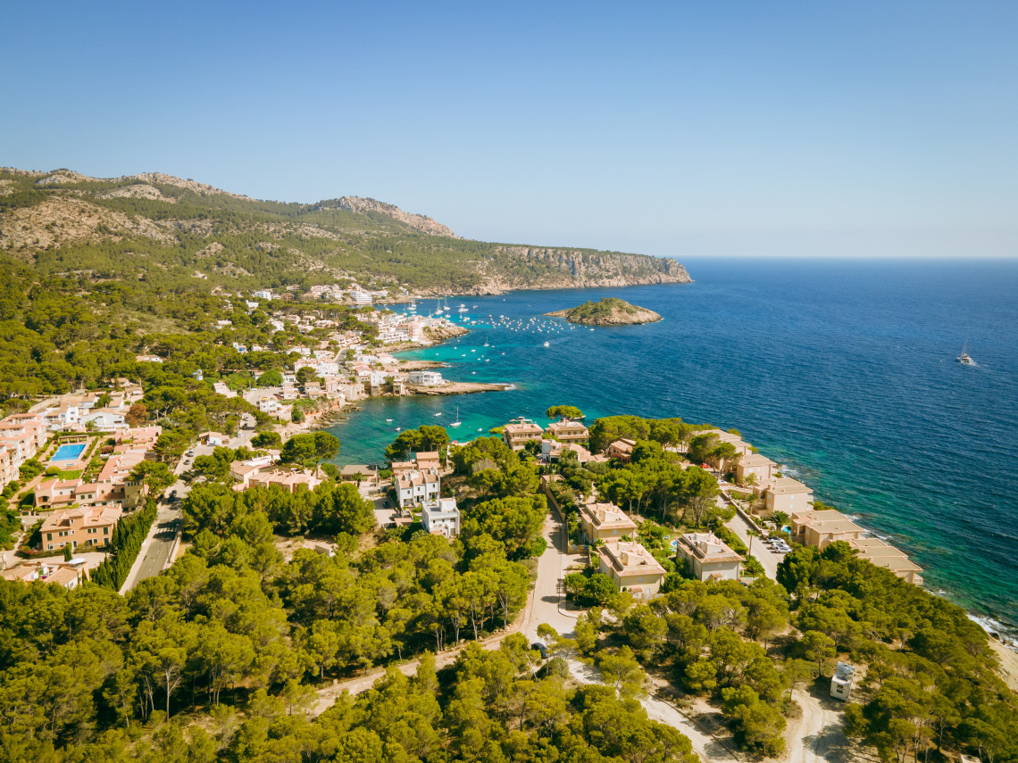 Aerial view of the Cala Fornells coastline in Calvi&agrave;, Spain, showing pine-covered hills, coastal villas, turquoise coves and boats anchored near a rocky islet off Mallorca.