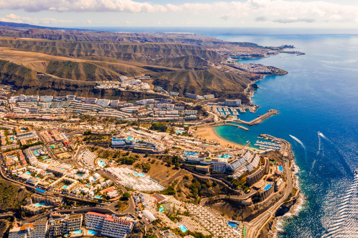 Aerial view of Puerto Rico in Mog&aacute;n, Spain, showing its sheltered beach, marina and resort buildings arranged between rugged hills and the Atlantic Ocean on Gran Canaria.