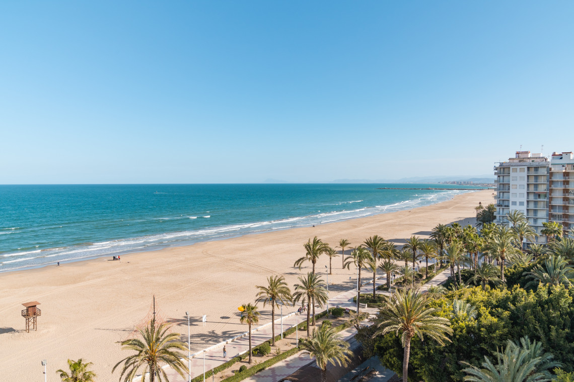 San Juan Beach in Alicante, Spain, extends along a wide sandy shoreline beside the Mediterranean Sea. Palm trees, a seafront promenade and residential buildings border the beach.
