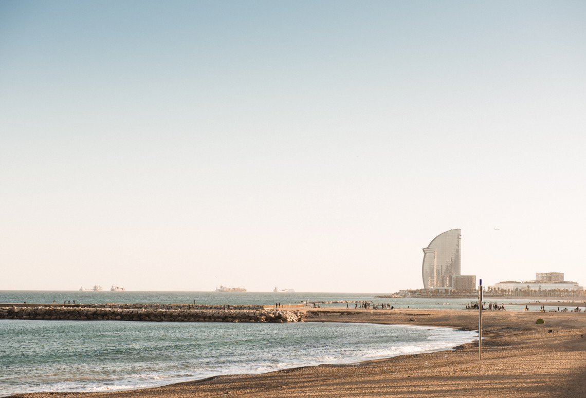 Sant Sebasti&agrave; Beach in Barcelona, Spain, stretches beside the Mediterranean Sea with the sail-shaped W Barcelona hotel rising along the waterfront. A stone breakwater separates the calm coastal water from the sandy beach