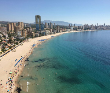 Aerial view of Levante Beach in Benidorm, Spain, showing the curved sandy shoreline, clear Mediterranean water and high-rise buildings extending along the coast.