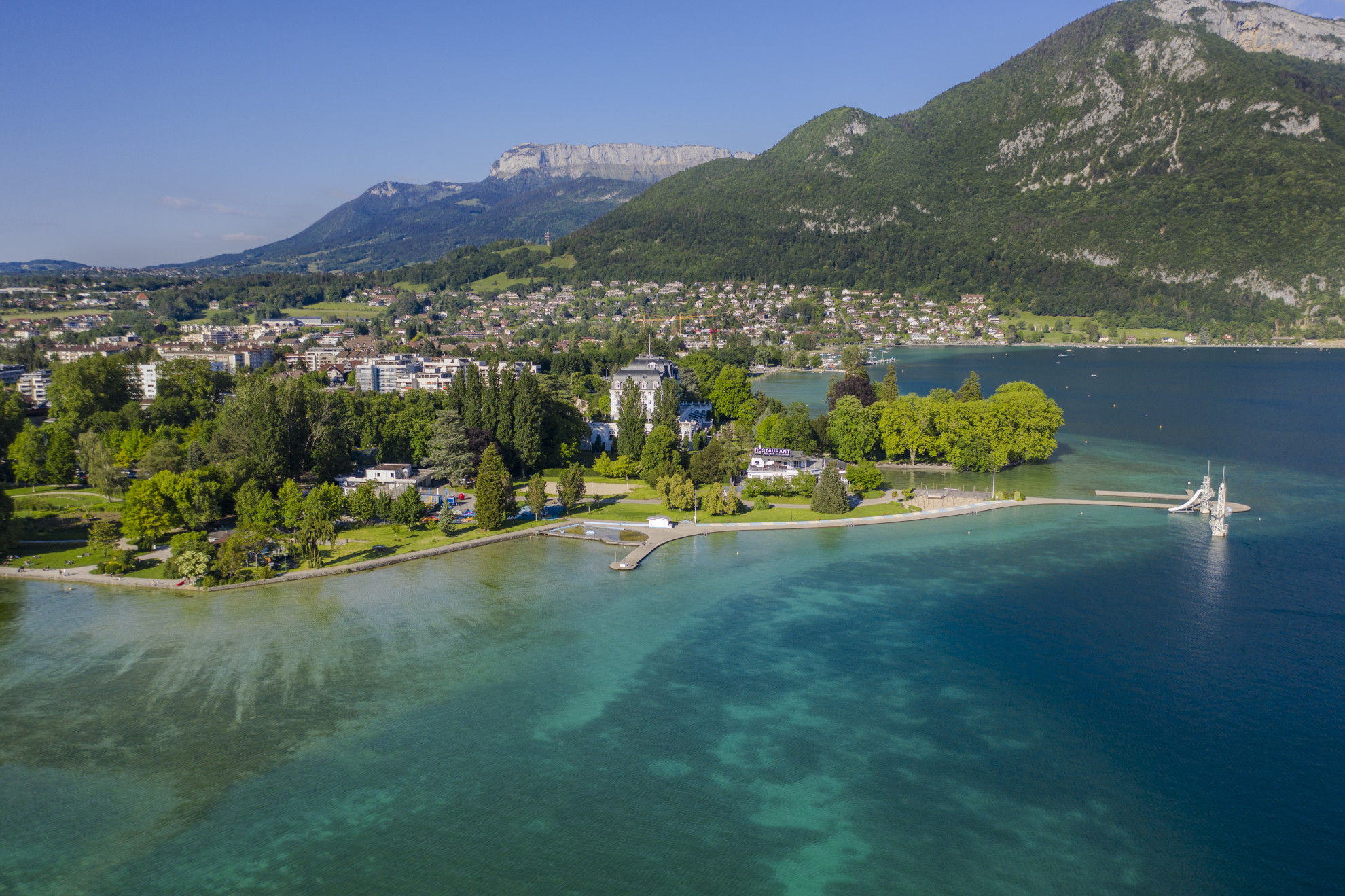 Aerial view of Lac d'Annecy with turquoise water, Belle Époque lakeside villa, ornate jetty, and limestone Alpine ridges, Haute-Savoie, France