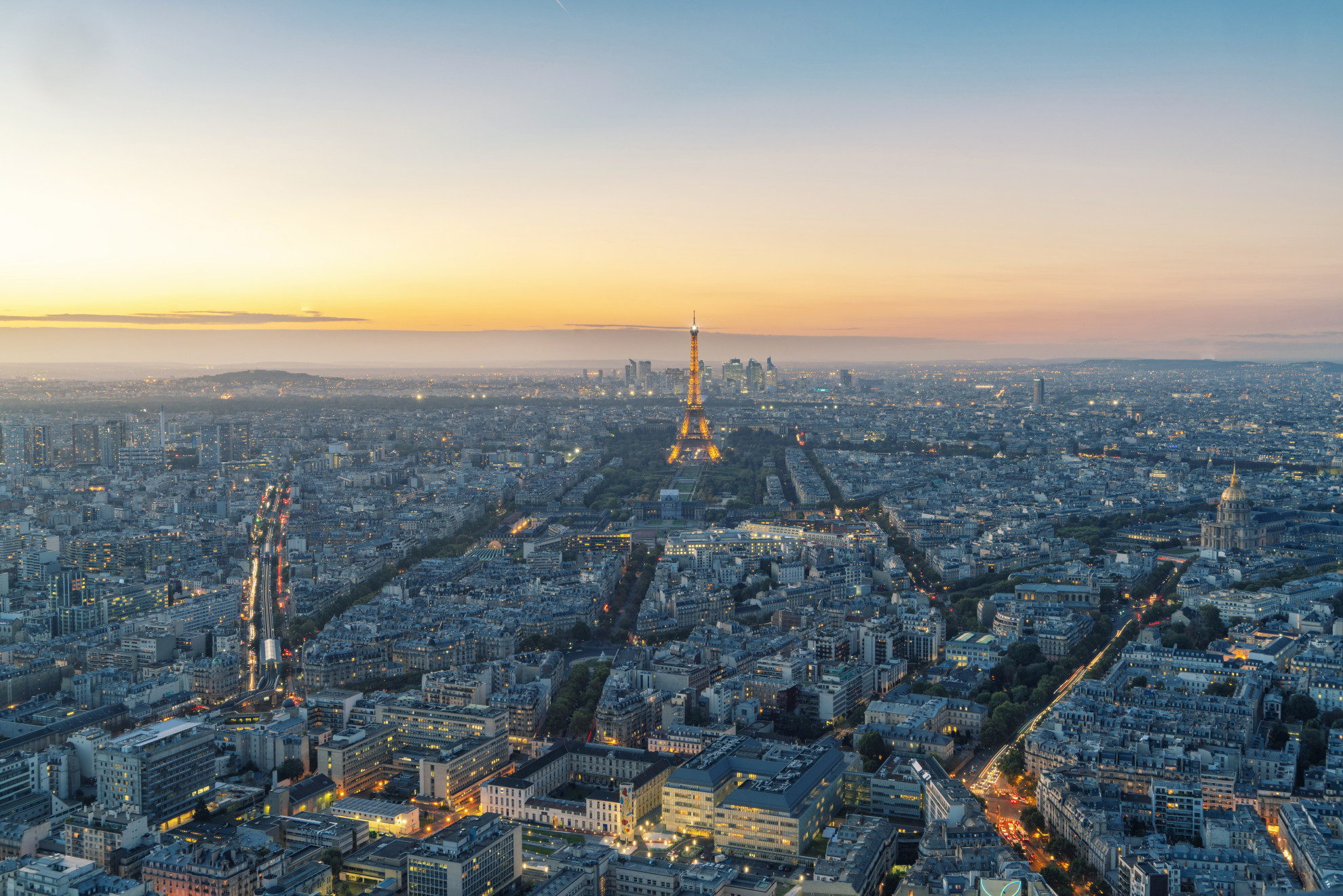 Aerial sunset view of Paris from above with the illuminated Eiffel Tower at centre, La Défense skyline in the distance, and the golden dome of Les Invalides visible to the right