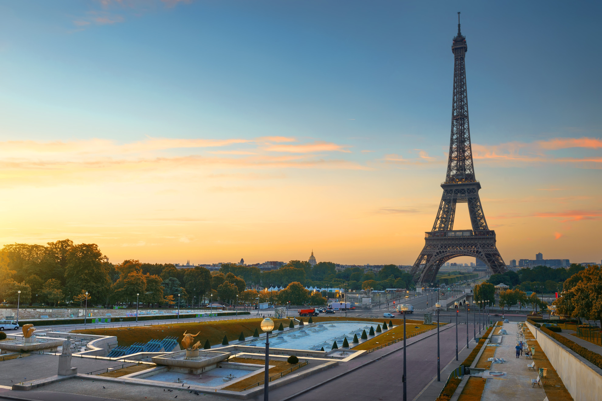 The Eiffel Tower at sunrise seen from the Trocadéro gardens, with golden morning light, fountains and esplanade in the foreground, and the dome of Les Invalides on the horizon