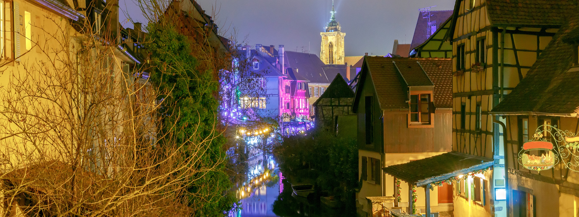 Evening view along Quai de la Poissonnerie in Colmar, France, with colourful Christmas lights reflected in the canal between traditional half-timbered houses.
