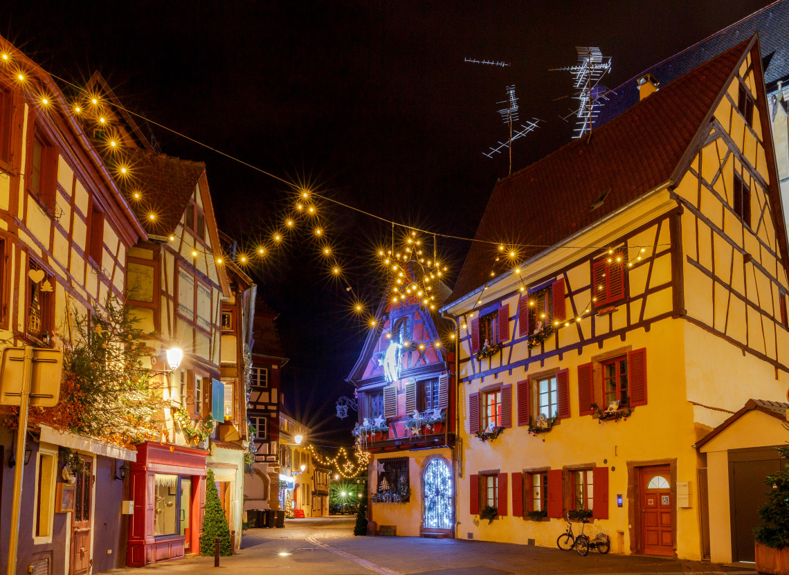 A quiet street in Colmar&rsquo;s historic old town, France, lined with traditional half-timbered houses and overhead Christmas lights at night.