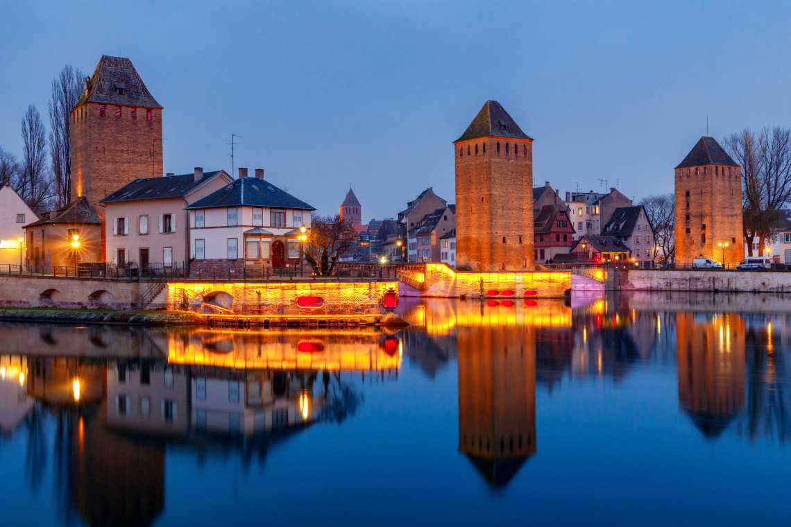 Evening view of the Ponts Couverts in Strasbourg, France, showing medieval defensive towers and bridges reflected in the calm waters of the River Ill.